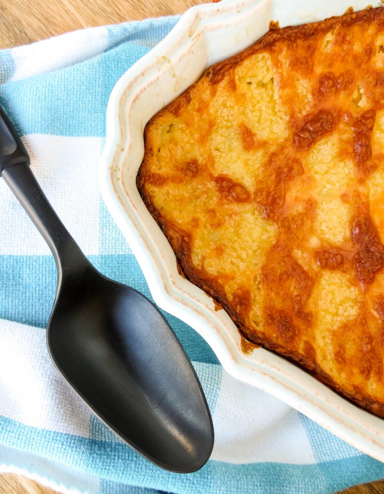 A casserole filled with Corn Casserole that has a browned topping of cheese with a black serving spoon on a blue and white towel.