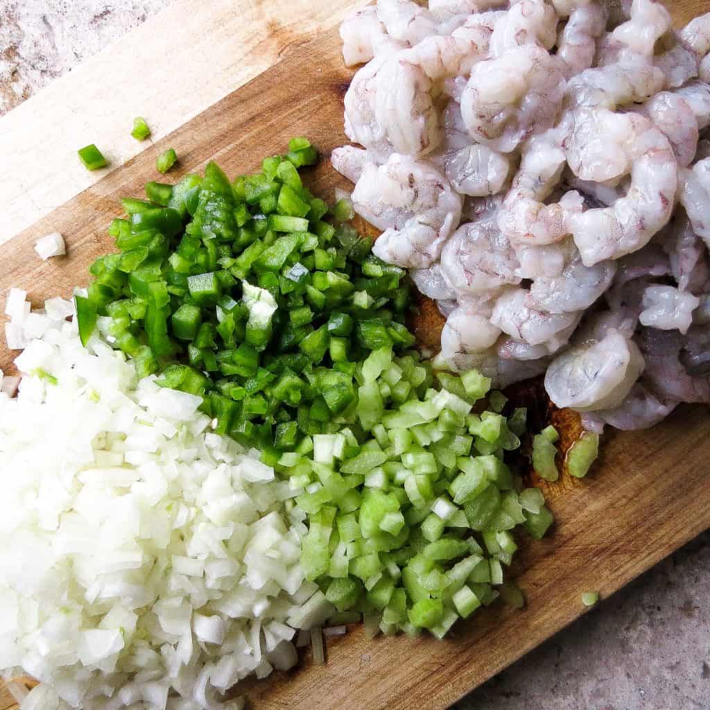 Chopped onions, green peppers, and celery on a wooden cutting board with whole peeled shrimp for Shrimp And Corn Soup.