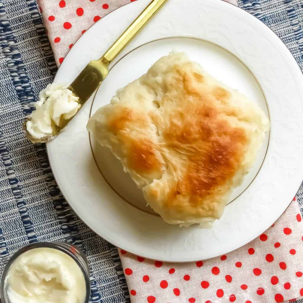An Easy No-Knead Bread roll on a white plate with a butter-slathered bread knife sitting on top of a white with red polka dotted napkin.