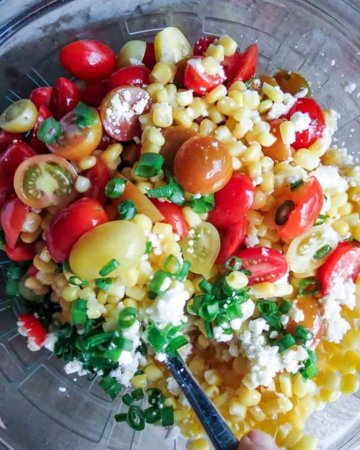 Glass bowl full of Corn Salad With Tomatoes stirred with a silver spoon.