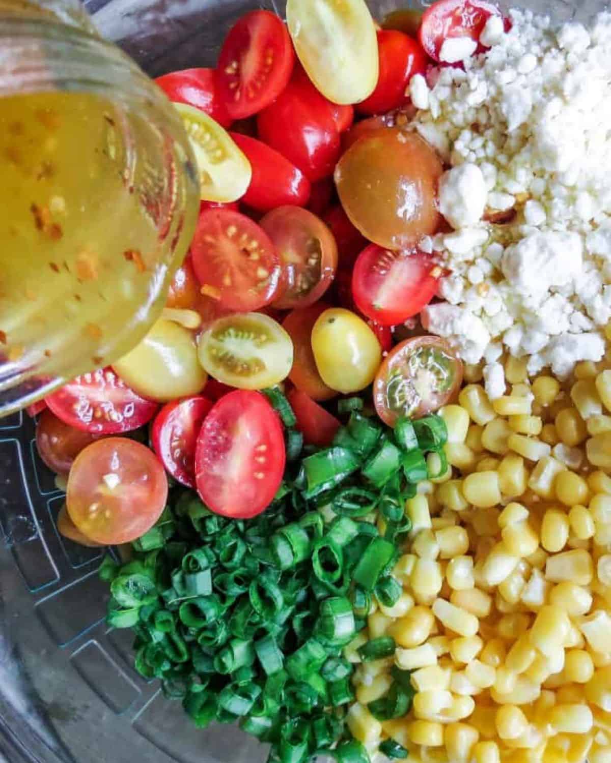 A glass bowl full of ingredients for Corn Salad With Tomatoes with a jar of vinaigrette being poured over it.