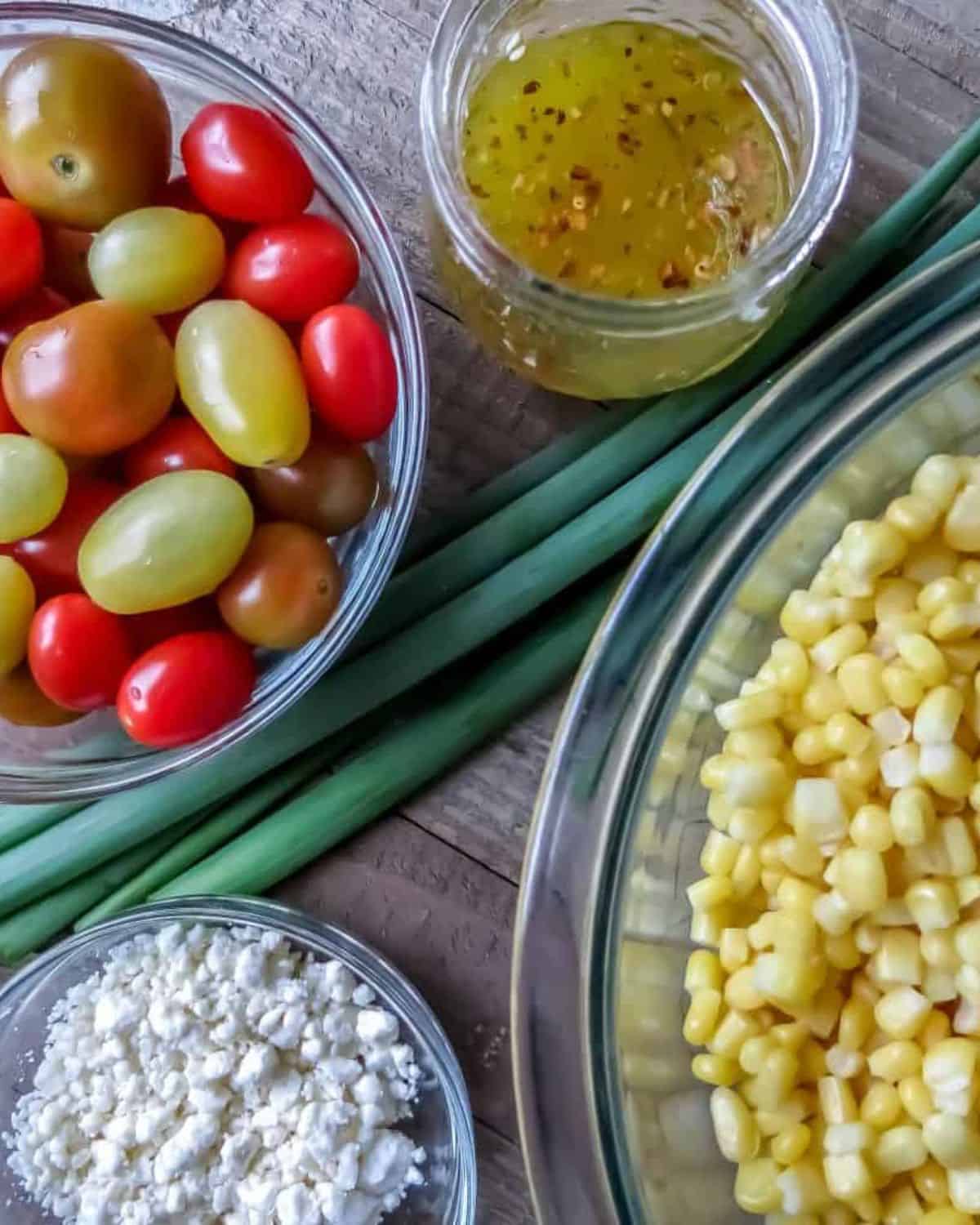 Individual bowls of corn, grape tomatoes, feta cheese, a jar of vinaigrette and some green onions on a wooden board for Corn Salad With Tomatoes.