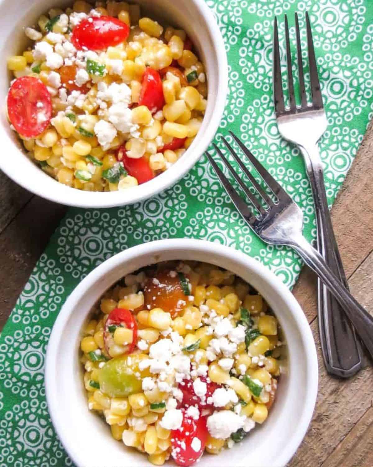 Two white bowls with each a serving of Corn Salad With Tomatoes on a green and white printed napkin with two forks.