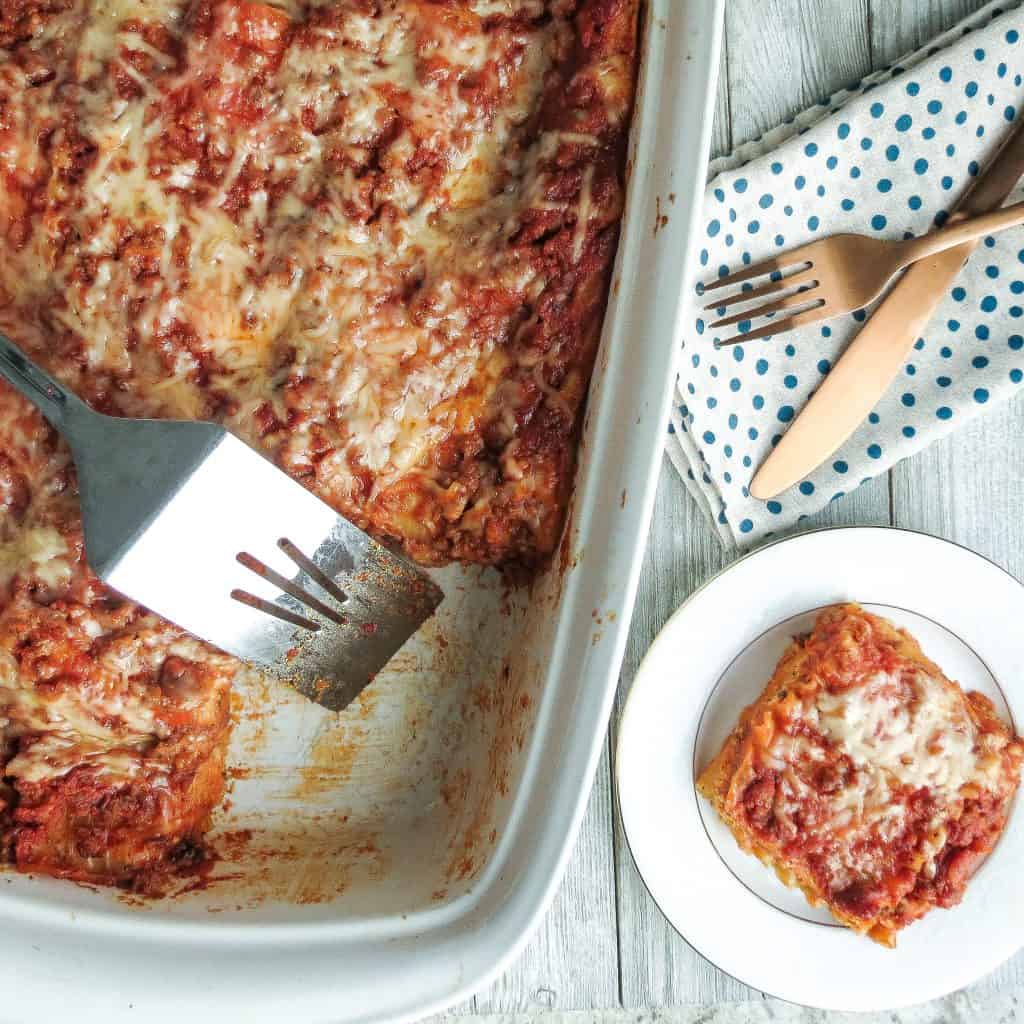 A white casserole dish of Easy Lasagna Recipe, Step-By-Step with a serving taken out and put on a white plate and a fork and knife on a blue and white napkin.