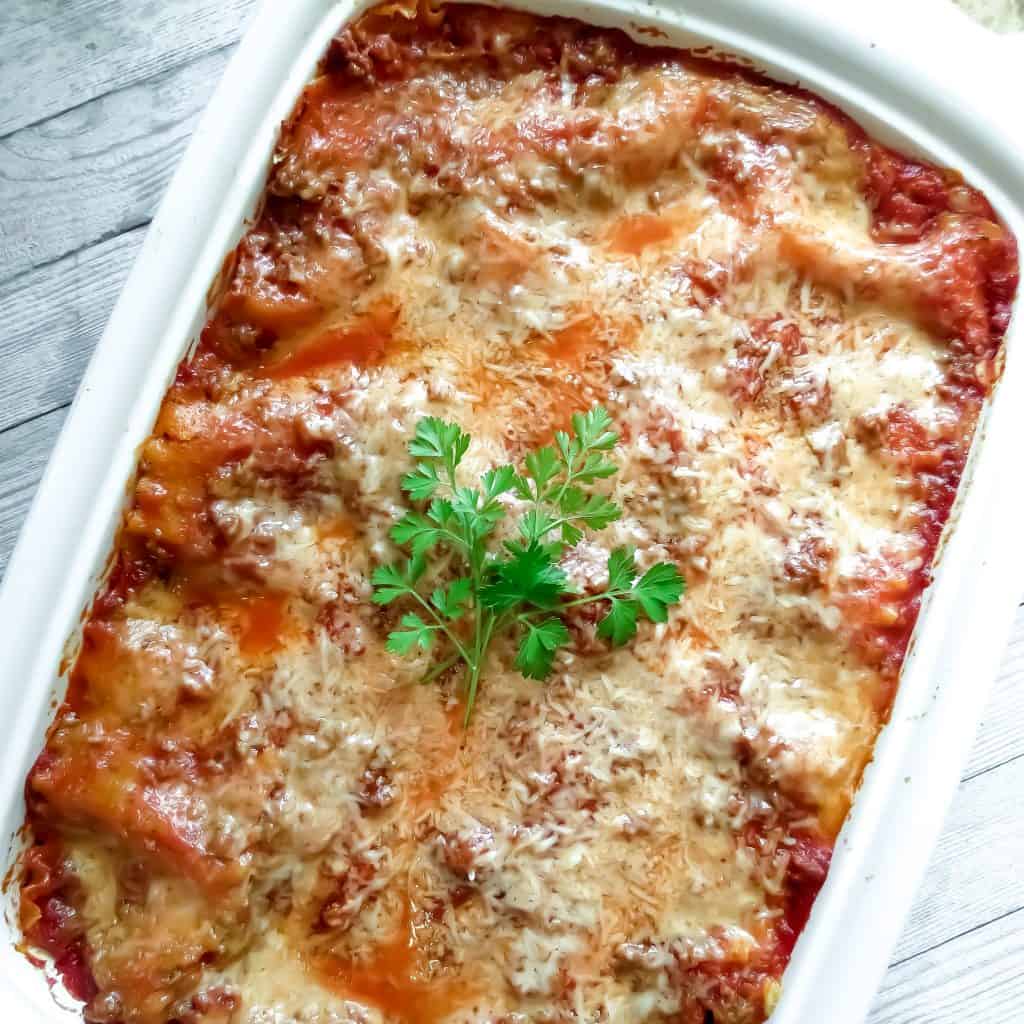 A white casserole dish with a cooked Easy Lasagna Recipe, Step-By-Step garnished with fresh parsley in the center.