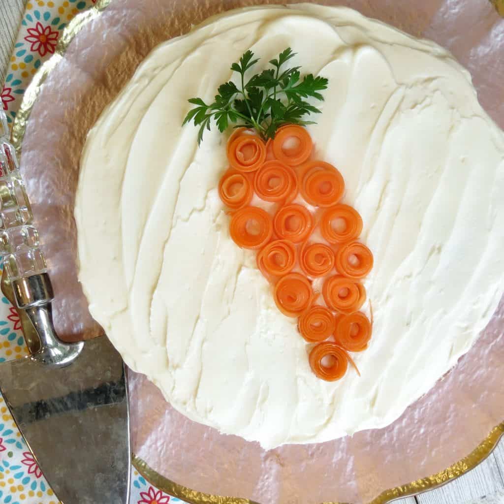 A Glazed Carrot Cake on a pink cake plate garnished with carrot curls and parsley leaves shaped to resemble a carrot.