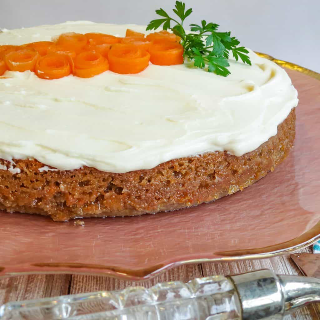 A Glazed Carrot Cake on a pink cake plate garnished with carrot curls and parsley leaves shaped to resemble a carrot.
