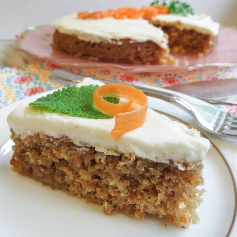 A slice of single layered Glazed Carrot Cake topped with a carrot curl and mint leaf on a white china plate.