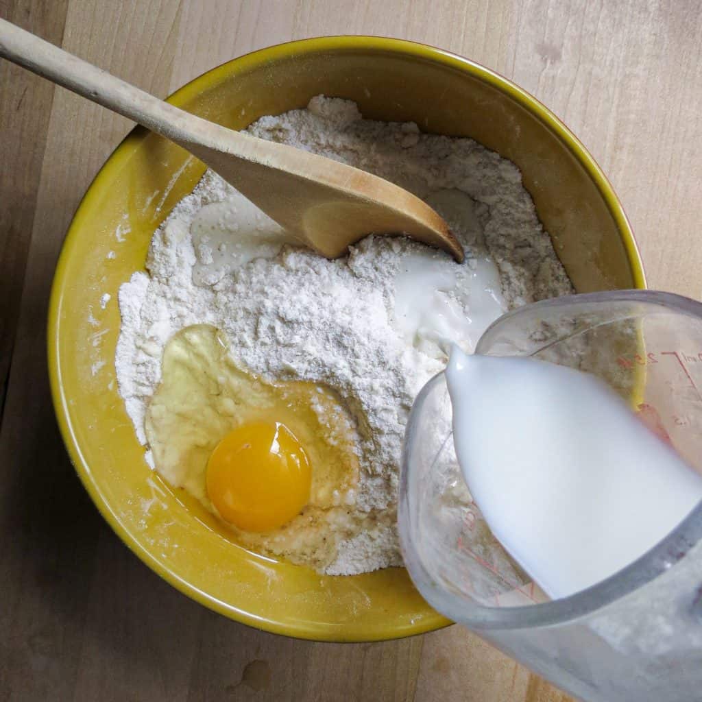A yellow bowl filled with flour and an egg with milk being poured into it.
