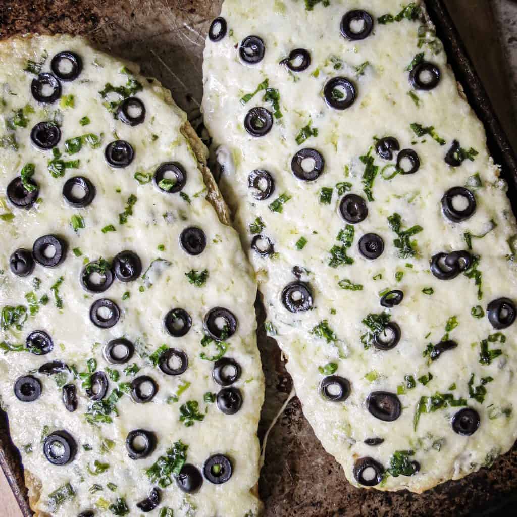 Two halves of French bread topped with melted cheese and ripe olives on a sheet pan.