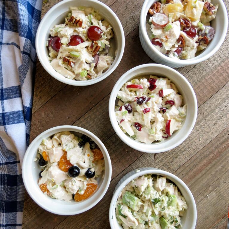 Five small white bowls on a wooden board of different chicken salads.
