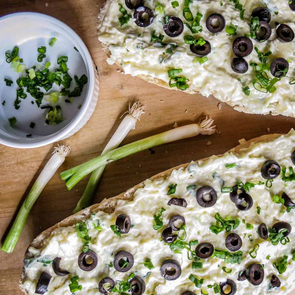 Chopped green onion tops in a white bowl next to 3 white rooted ends of green onions next to Cheesy Greek Bread on a wooden board.