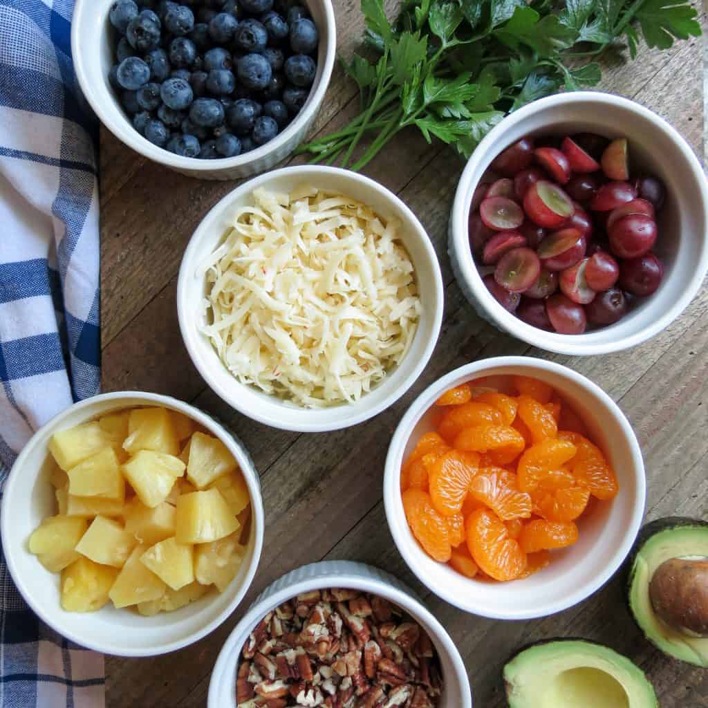 Six white bowls with fruit, nuts, and cheese on a wooden board with parsley and a cut avocado.