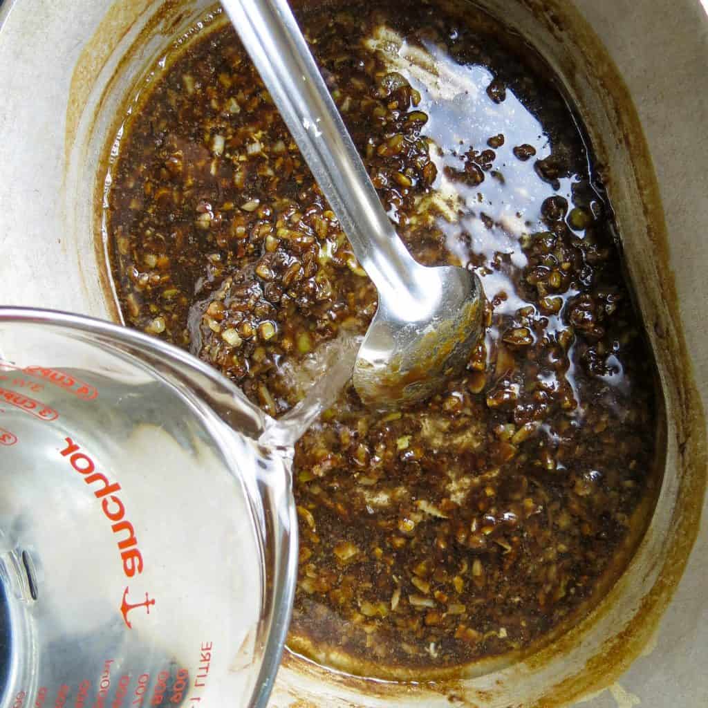 A measuring cup of water poured into silver pot of roux stirred with a silver spoon.