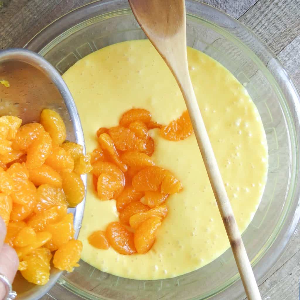 Mandarin oranges poured into a yellow cake batter with a wooden spoon resting on the glass bowl.