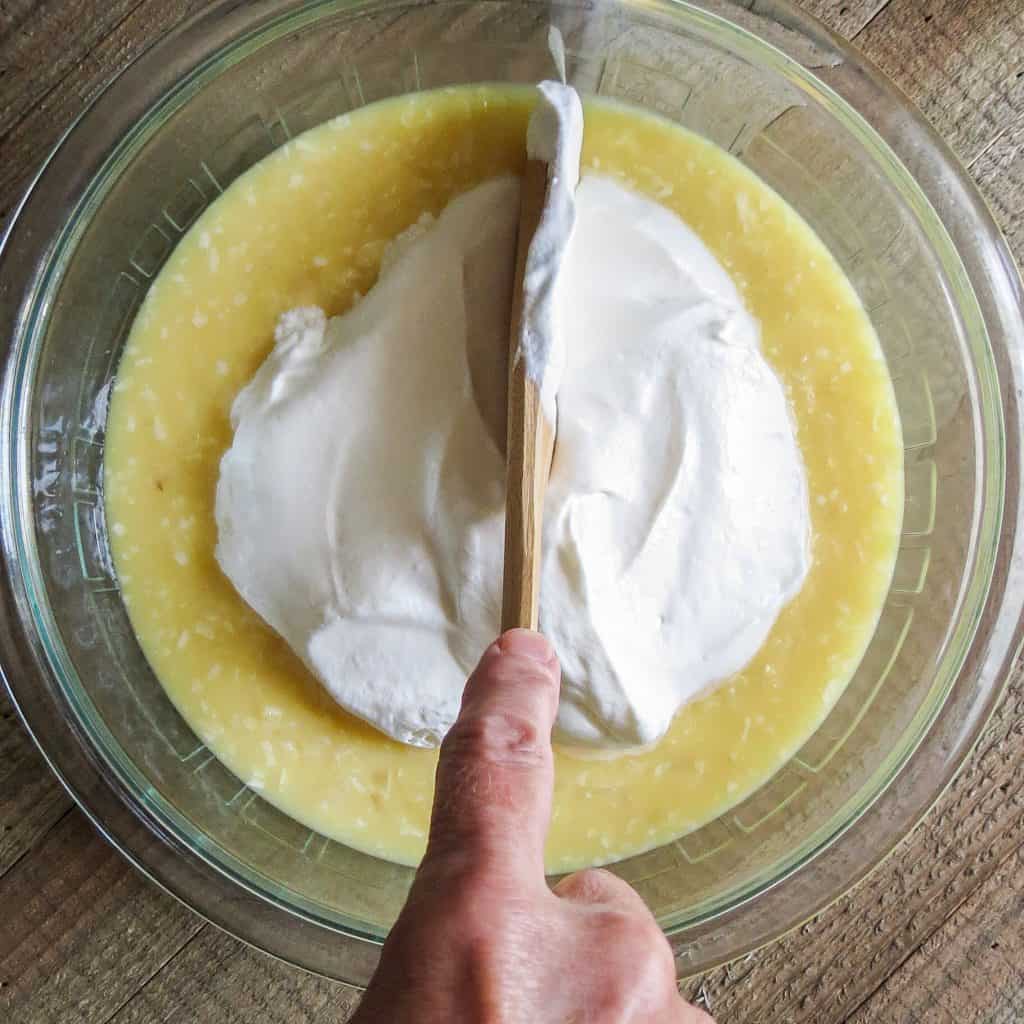 A glass bowl of coconut pudding with whipped cream on top stirred with a wooden spoon.