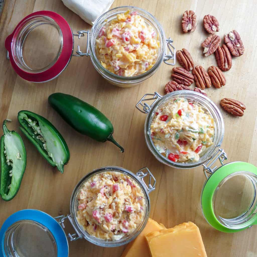 Three pimento cheeses in glass containers with jalapeño peppers, cheese, and pecans on a wooden board.