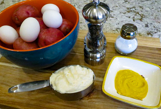 A bowl of fresh eggs and potatoes, a cup of mayonnaise, a bowl of mustard, a pepper mill, and a salt shaker on a wooden cutting board.