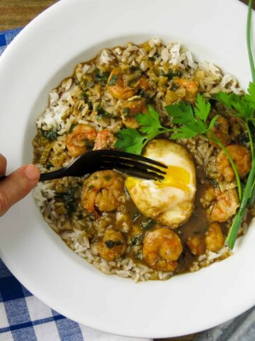 A white bowl of shrimp stew on rice with a poached eggs cut into with a fork.