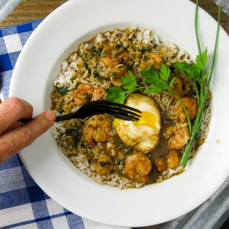 A white bowl of shrimp stew on rice with a poached eggs cut into with a fork.
