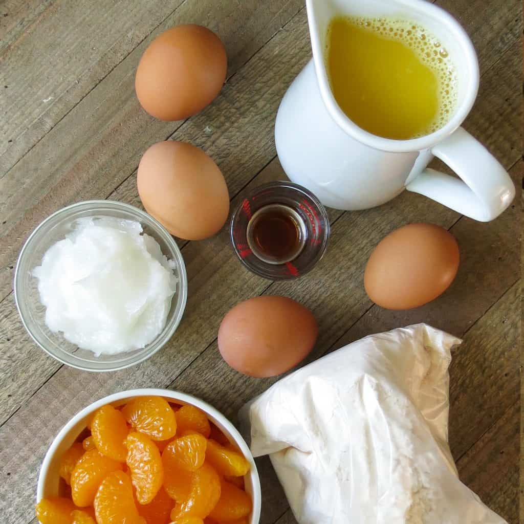 Four eggs, a white pitcher of orange juice, a bowl of mandarin oranges, bag of cake batter, a bowl of coconut oil, and vanilla flavoring on a wooden bowl.