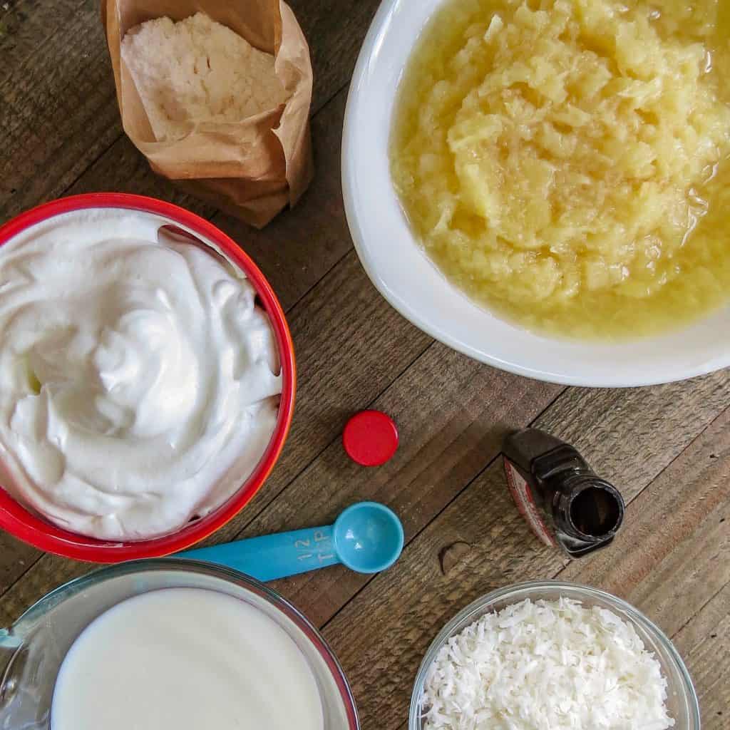 A white bowl of crushed pineapple, package of instant pudding, red bowl of whipped cream, coconut, measuring spoon and bottle of flavoring, and pitcher of milk on a wooden board.