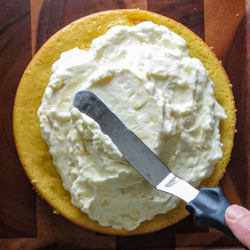 A round yellow cake layer frosted with a creamy filling with a slanted spatula on a wooden board.