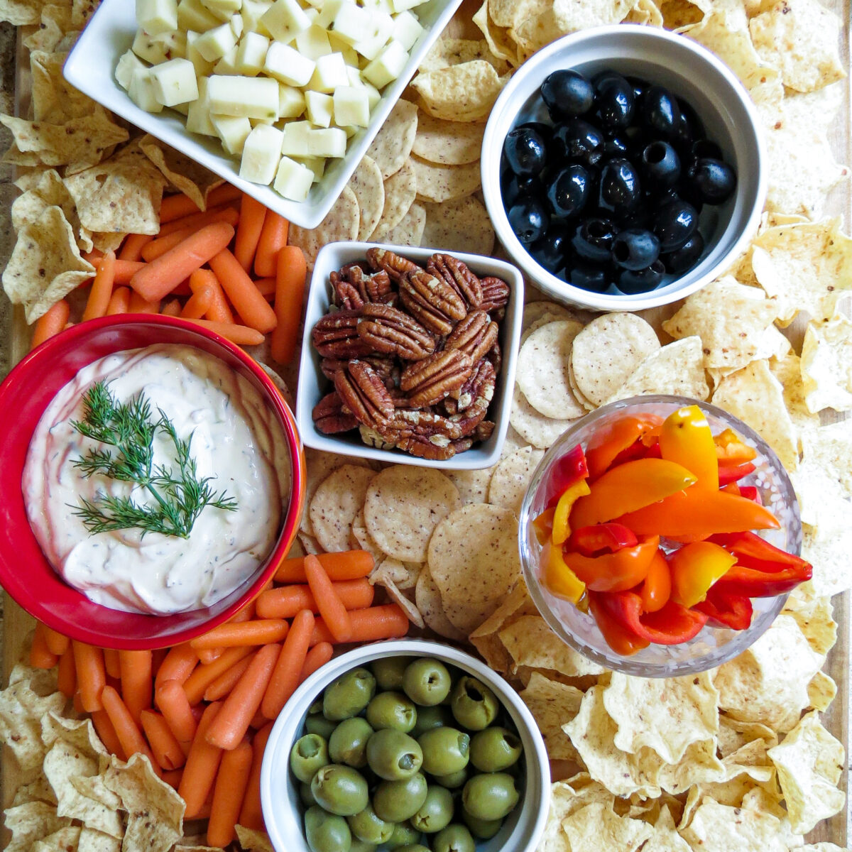 A board filled with white bowls filled with snacks with crackers surrounding them.