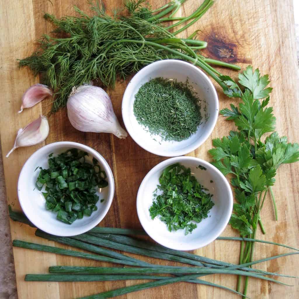 A wooden board with fresh herbs and a bulb of garlic.