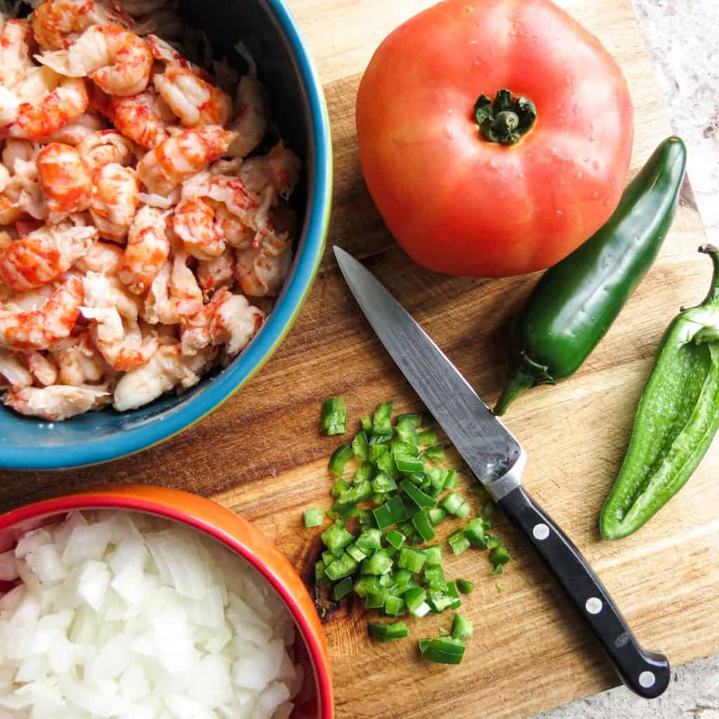 Ingredients for Crawfish 'n Cornbread of jalapeño peppers, tomato, onions, and crawfish tails on a wooden board with a knife.