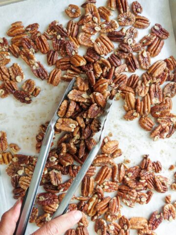 Pecan halves spread on a parchment paper lined pan with tongs grabbing pecans.