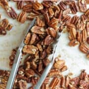 Pecan halves spread on a parchment paper lined pan with tongs grabbing pecans.