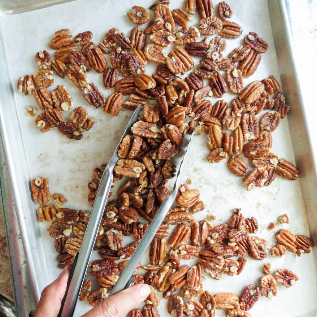 Pecans on a sheet pan lined with parchment paper tossed with long-handled tongs.