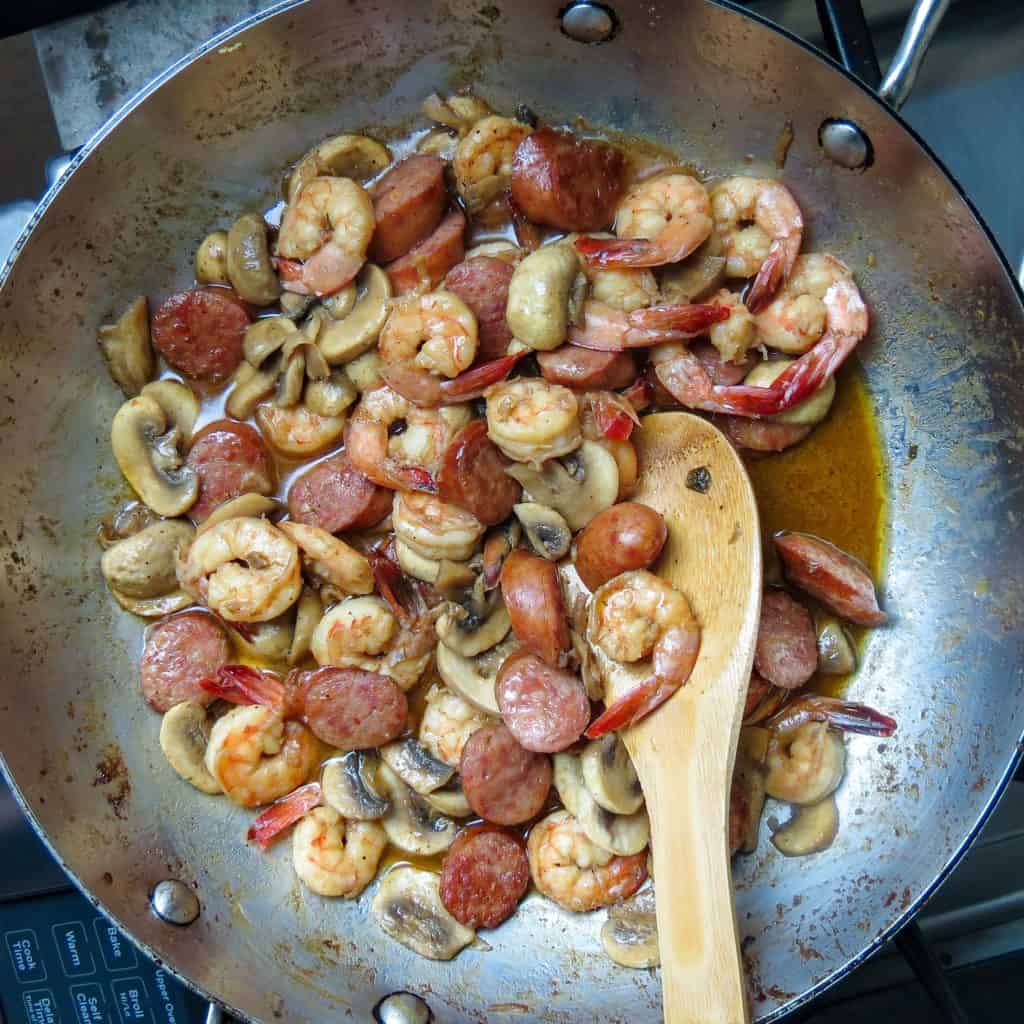 Cooked shrimp, sausage, and mushrooms in a spicy sauce in a pan stirred with a wooden spoon.