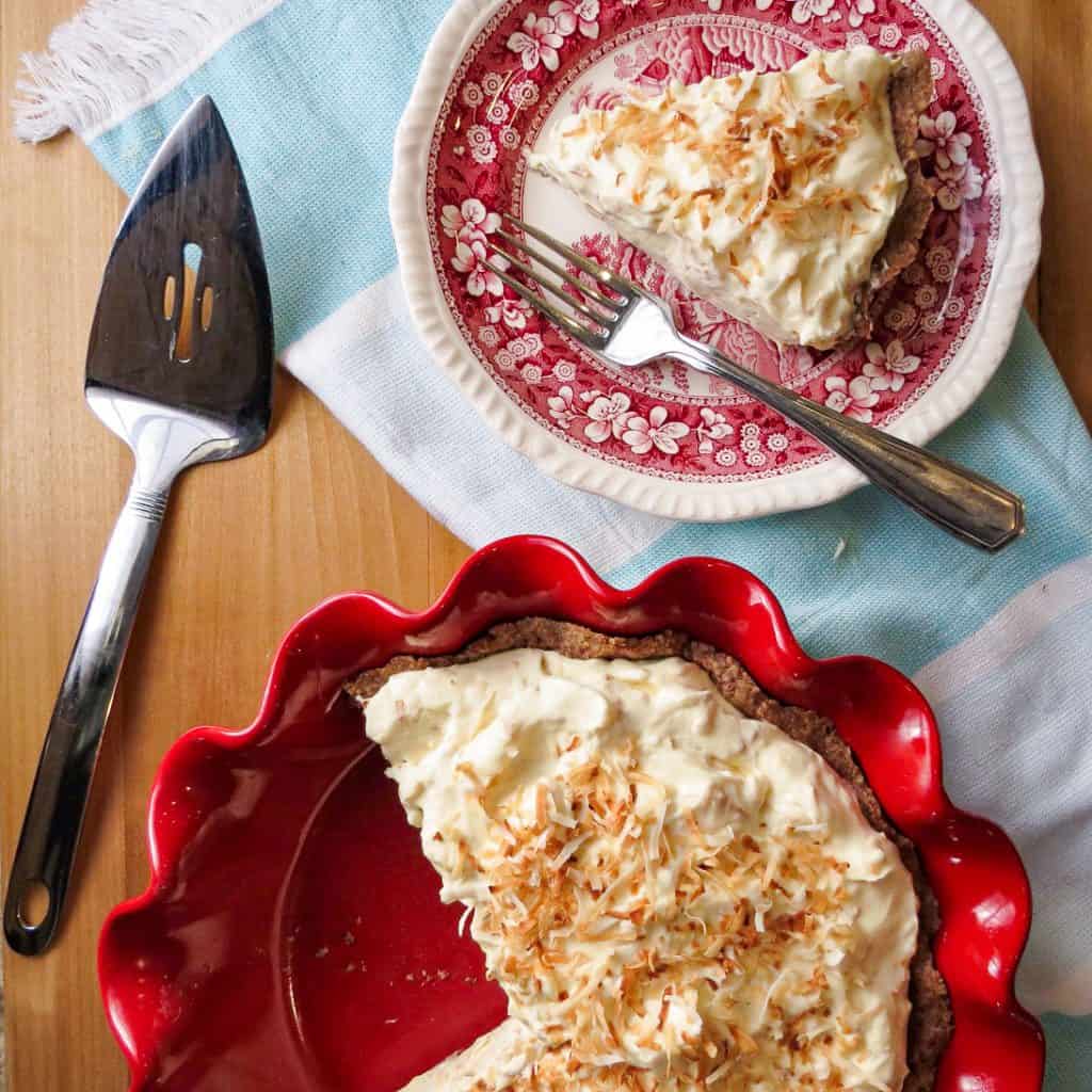 A piece of Low Carb Coconut Pie on a plate, a pie in a red pie pan with a pie server.