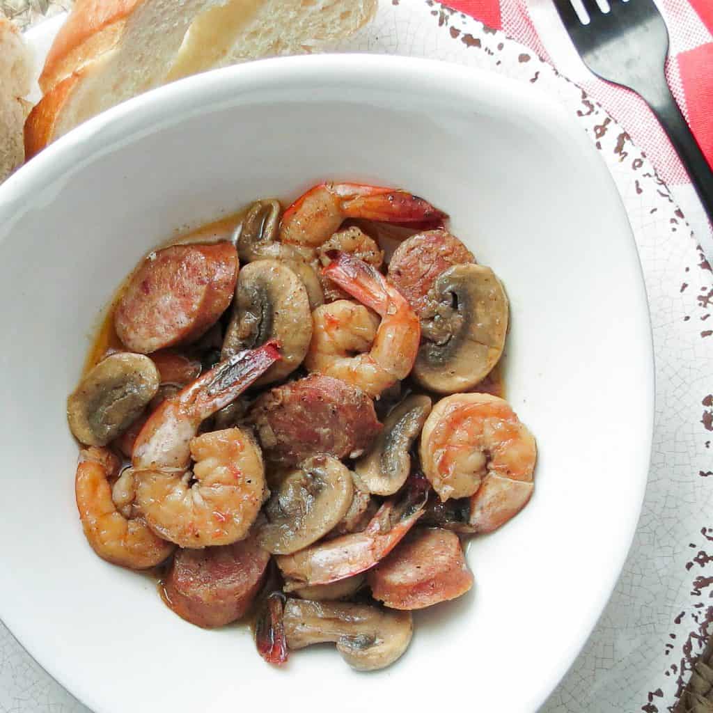 A bowl full of barbecue shrimp with mushrooms and smoked sausage.