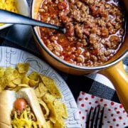 Chili in a pot next to a plate of chili dog and chips.