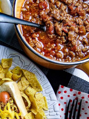 Chili in a pot next to a plate of chili dog and chips.