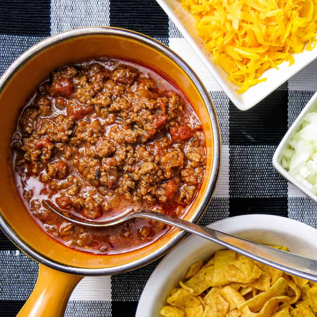 A pot of chili next to bowls of cheese, onions, and chips.