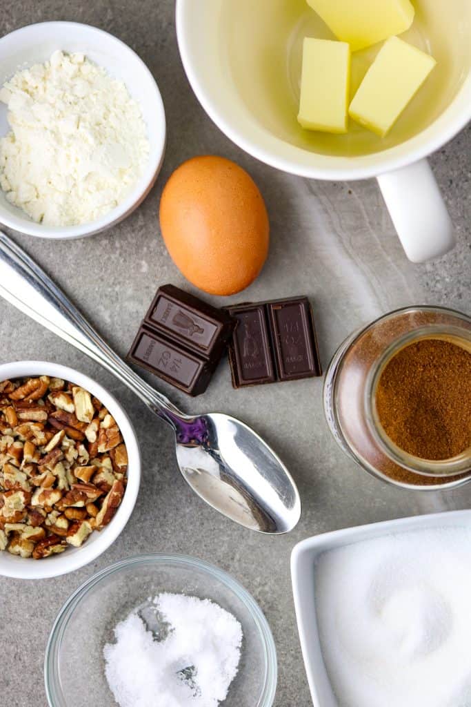 Baking ingredients with chocolate squares, nuts, egg, and butter spread out on a gray countertop.