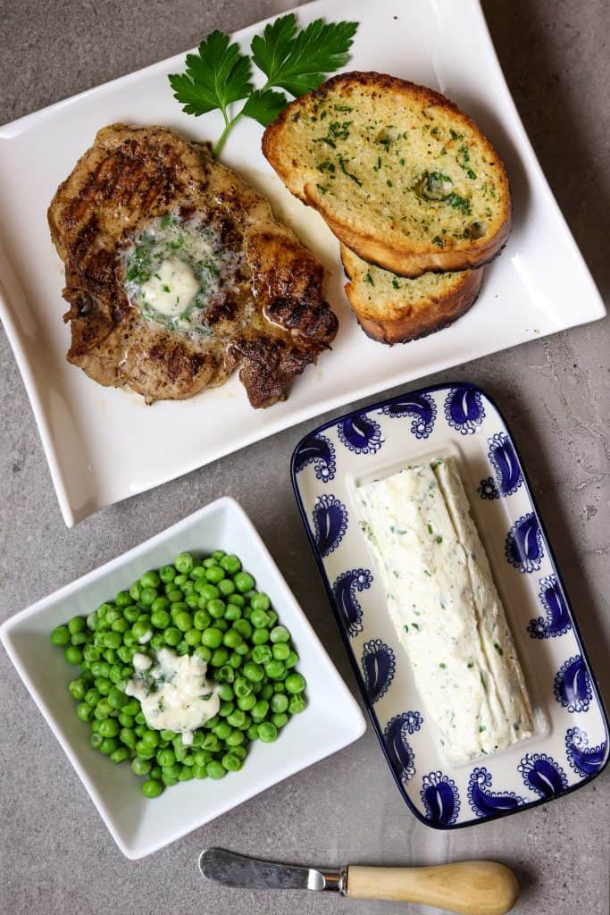 A log of Herb Butter with a plate of bread and a pork chop with a side of green peas.