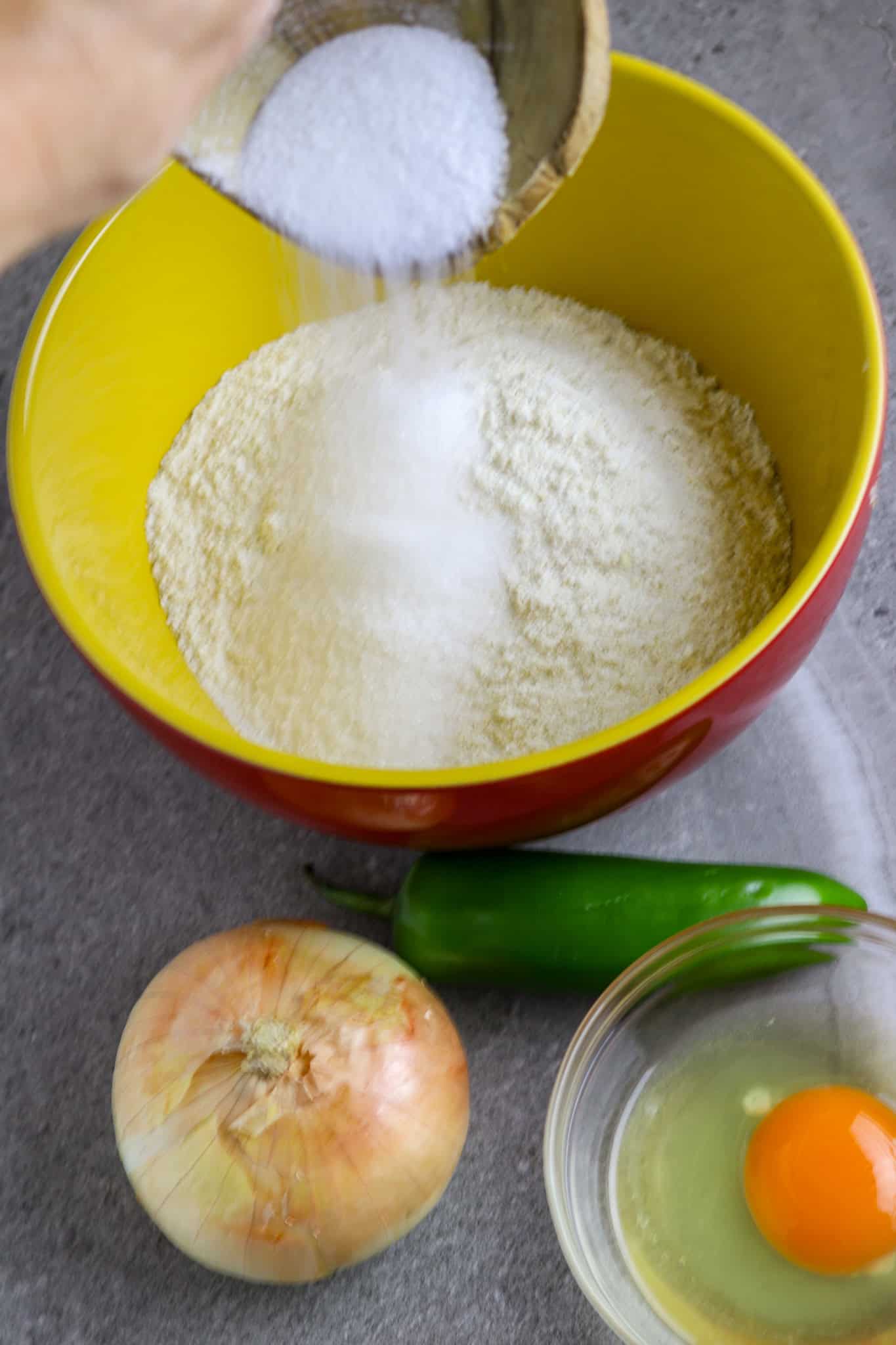 A medium bowl of corn meal mix with sugar, a whole onion, and jalapeno pepper with an egg in a bowl next to the medium bowl.