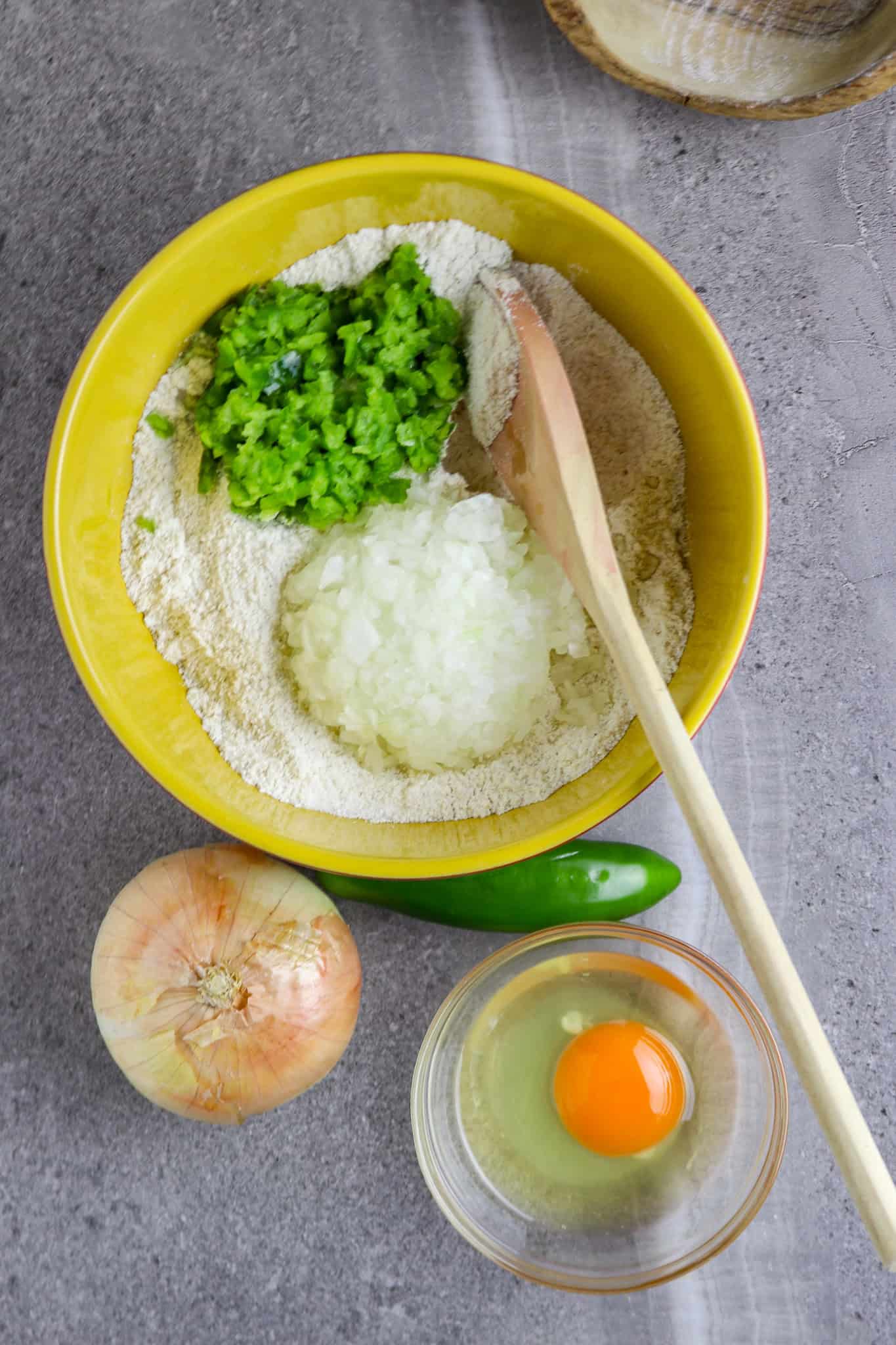 A medium bowl of corn meal mix with chopped jalapeno pepper and onion in the bowl with an egg in a small bowl next to the medium bowl with a whole onion and pepper next to it.
