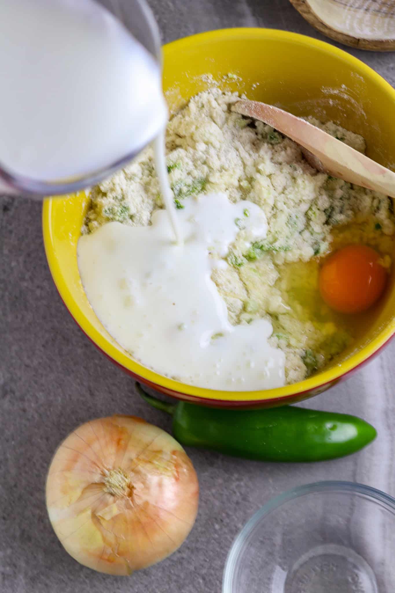 A bowl of corn meal mix with an egg and pitcher of buttermilk being poured into the bowl.