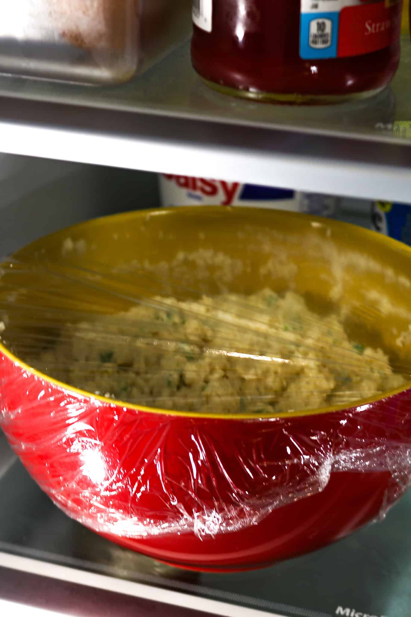 A red bowl with hush puppy mix in it and covered with cellophane over it in the refrigerator.