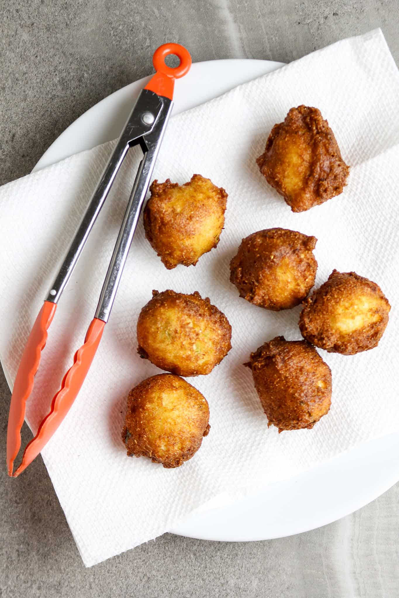 Golden fried hush puppies draining on a white paper towel with a pair of orange handled tongs.