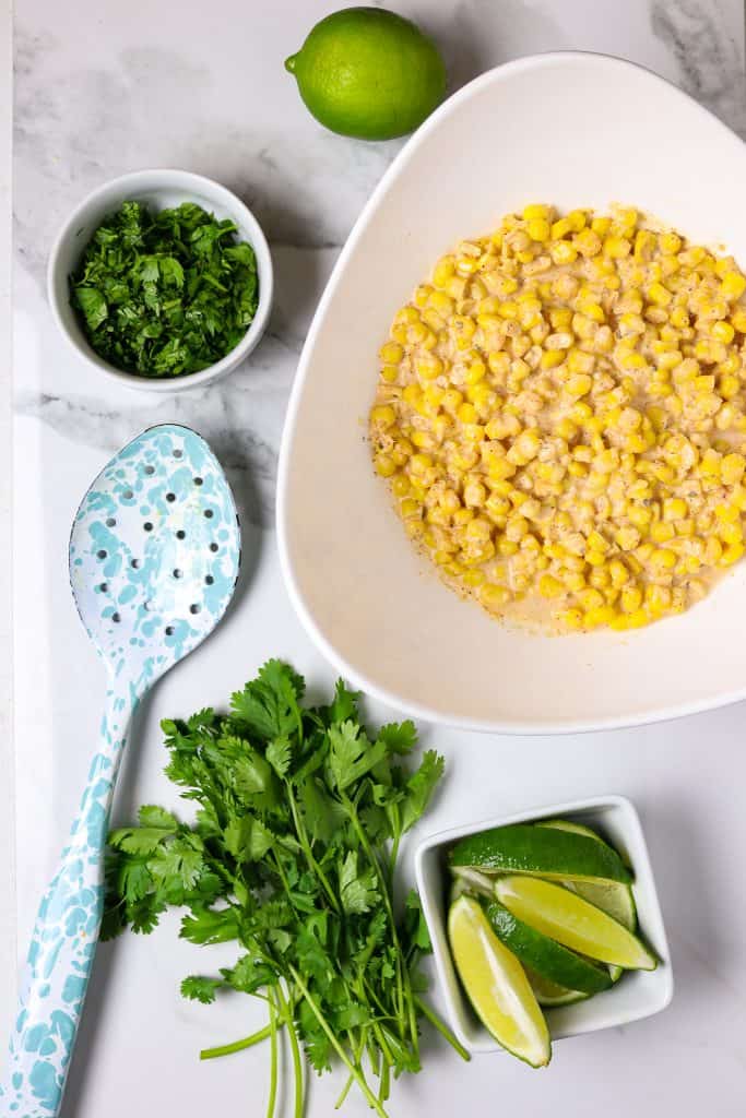 A bowl of corn with a bowl of limes and sprigs of cilantro and a serving spoon.