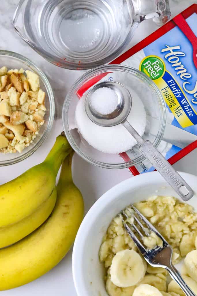 Mashed bananas in a bowl with a fork, a bowl of crushed vanilla wafers and a tablespoon of sugar.