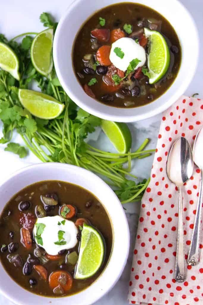 Two white bowls of Black Bean soup with lime, cilantro, and sour cream to garnish.