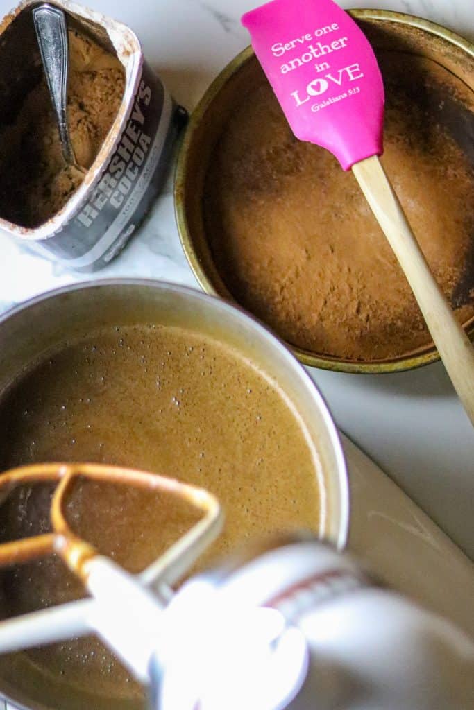 A mixing bowl of batter with a spatula resting on a baking pan near a can of cocoa powder.